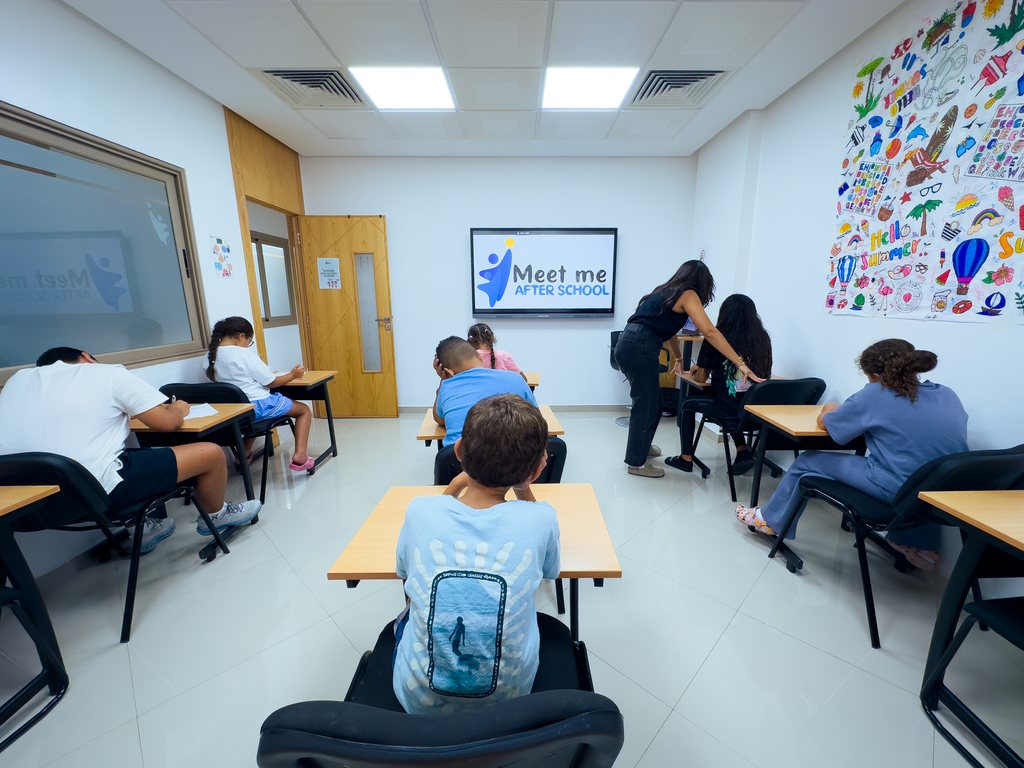 Des enfants installés à des tables individuelles dans une salle de classe, sous la supervision d'une adulte, face à un écran affichant « Meet me after school ».